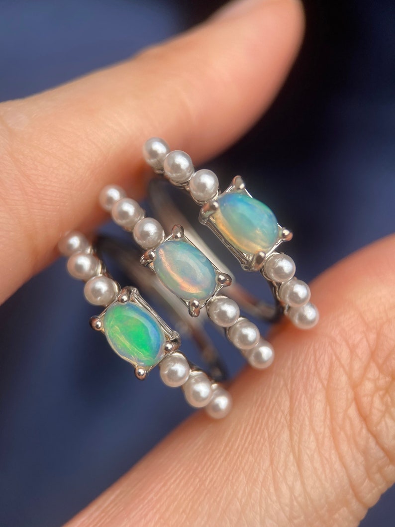 May include: Three silver rings with opal gemstones and white pearl accents. The rings are stacked on a finger.