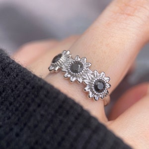 May include: A silver ring with three black gemstones set in sunflower designs.