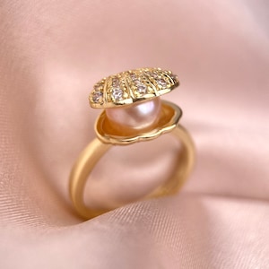 May include: A gold ring with a pearl inside a shell-shaped setting. The shell is decorated with small clear stones.