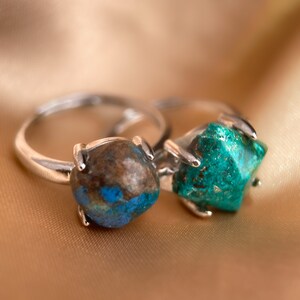 May include: Two silver rings with green and brown gemstone settings. The rings are on a gold fabric background.