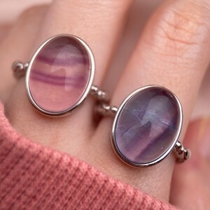 May include: Two silver rings with oval-shaped purple and pink fluorite gemstones. The rings are on a finger.