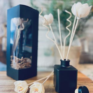 Puede incluir: Un difusor cuadrado negro con varillas de flores blancas y una tapa negra. Dos varillas de flores blancas están sobre una superficie de madera. Una caja negra con una ventana transparente está en el fondo.