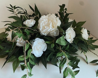 May include: A white floral arrangement featuring white flowers and green foliage. The flowers are arranged in a cascading style, with the leaves trailing down the sides.