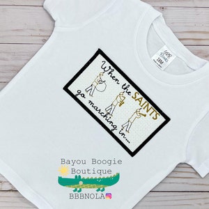 Peut inclure: T-shirt blanc &agrave; manches courtes avec un motif brod&eacute;. Le motif comprend le texte "When the SAINTS go marching in..." et des illustrations de musiciens. Le t-shirt pr&eacute;sente &eacute;galement le logo "Bayou Boogie Boutique" et un alligator vert.