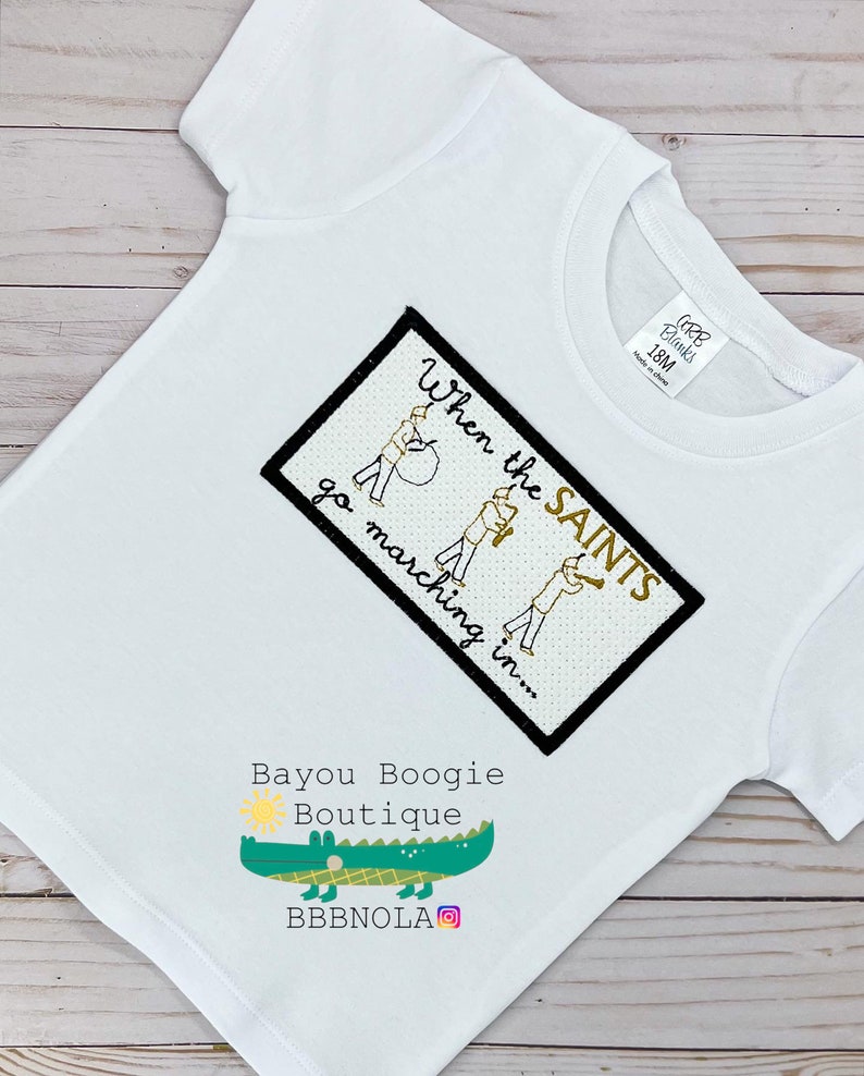 Peut inclure: T-shirt blanc &agrave; manches courtes avec un motif brod&eacute;. Le motif comprend le texte "When the SAINTS go marching in..." et des illustrations de musiciens. Le t-shirt pr&eacute;sente &eacute;galement le logo "Bayou Boogie Boutique" et un alligator vert.