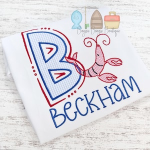 May include: White t-shirt with a large, embroidered blue and red letter "B" with a gingham pattern. A red embroidered lobster and the name "Beckham" in blue complete the design. The shirt has a fishing theme.