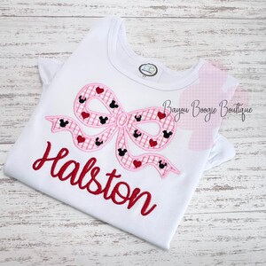 May include: White t-shirt with a pink bow design featuring Mickey Mouse silhouettes and red hearts. The name "Halston" is embroidered in red script below the bow. A small tag is visible near the neckline.
