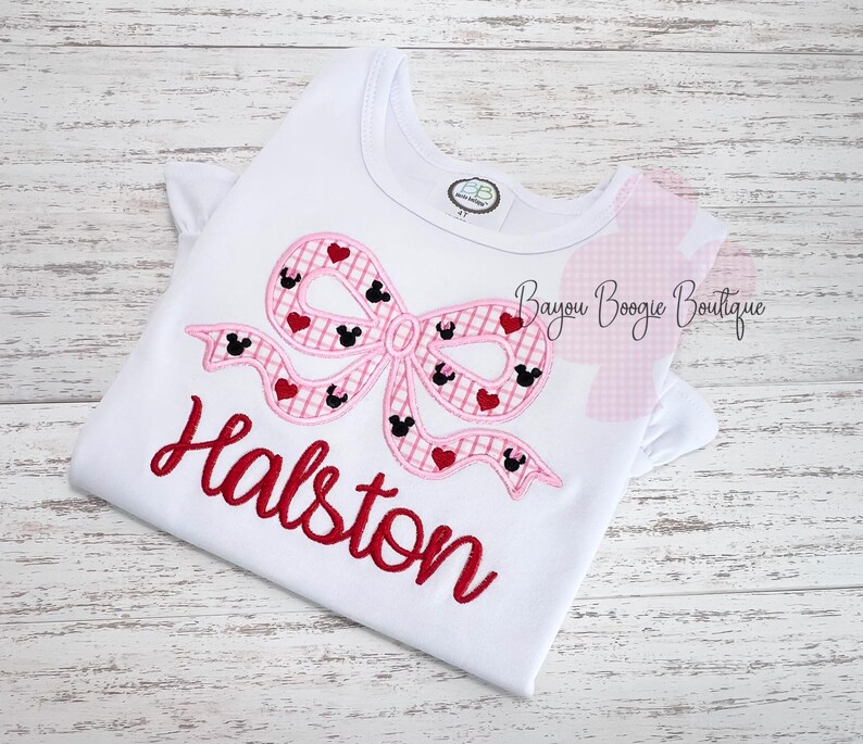 May include: White t-shirt with a pink bow design featuring Mickey Mouse silhouettes and red hearts. The name "Halston" is embroidered in red script below the bow. A small tag is visible near the neckline.