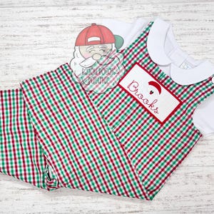 May include: A Christmas-themed outfit featuring a white collared shirt and a red, green, and white checkered overall. The overall has a rectangular patch embroidered with the name "Brooks" and a Santa Claus design. A Santa Claus graphic is also visible.