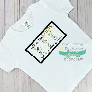Peut inclure: T-shirt blanc &agrave; manches courtes avec un motif brod&eacute;. Le motif pr&eacute;sente le texte "When the SAINTS go marching in..." et des illustrations de membres d'un groupe de musique. Le t-shirt porte &eacute;galement le logo Bayou Boogie Boutique.