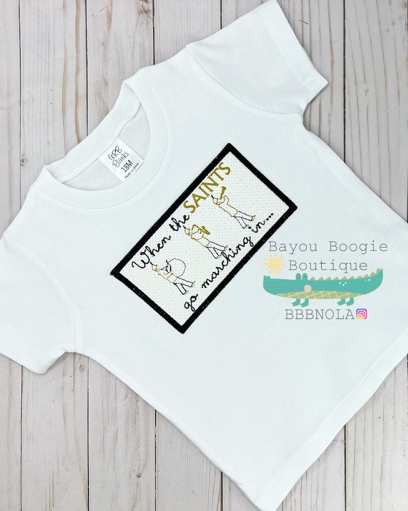Peut inclure: T-shirt blanc &agrave; manches courtes avec un motif brod&eacute;. Le motif pr&eacute;sente le texte "When the SAINTS go marching in..." et des illustrations de membres d'un groupe de musique. Le t-shirt porte &eacute;galement le logo Bayou Boogie Boutique.