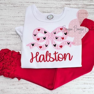 May include: A white t-shirt and red pants set. The shirt features a pink bow design with Mickey Mouse silhouettes and hearts, with the name "Halston" embroidered in red. A red floral accent is on the left.