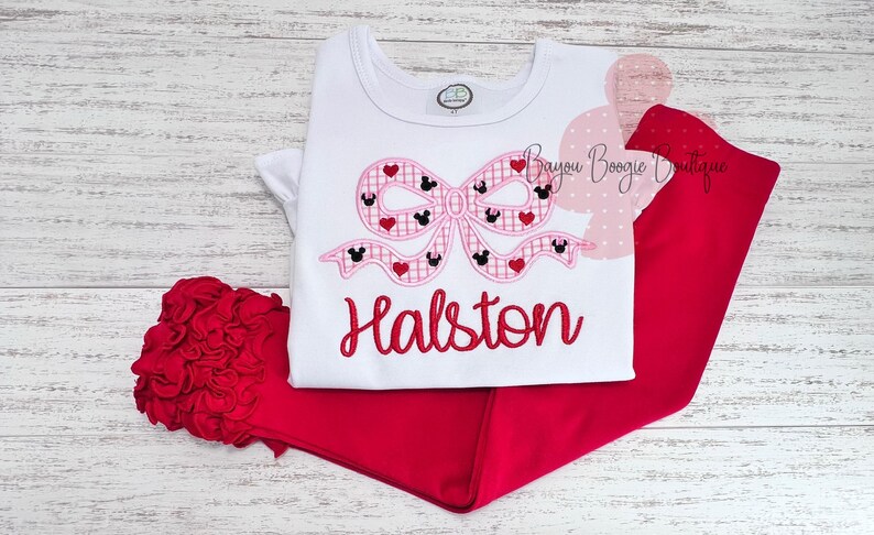 May include: A white t-shirt and red pants set. The shirt features a pink bow design with Mickey Mouse silhouettes and hearts, with the name "Halston" embroidered in red. A red floral accent is on the left.