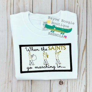 Peut inclure: T-shirt blanc &agrave; col rond avec un motif brod&eacute; noir et or. Le motif pr&eacute;sente le texte "When the SAINTS go marching in..." et des illustrations de musiciens. Une &eacute;tiquette avec un logo d'alligator vert est attach&eacute;e.