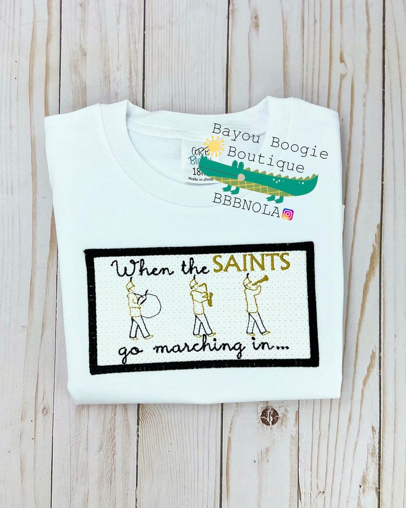 Peut inclure: T-shirt blanc &agrave; col rond avec un motif brod&eacute; noir et or. Le motif pr&eacute;sente le texte "When the SAINTS go marching in..." et des illustrations de musiciens. Une &eacute;tiquette avec un logo d'alligator vert est attach&eacute;e.