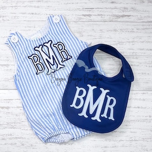 May include: A baby romper and bib set. The romper is light blue and white striped with a monogram. The bib is navy blue with a matching monogram. The text "Bayou Boogie Boutique" is also visible.