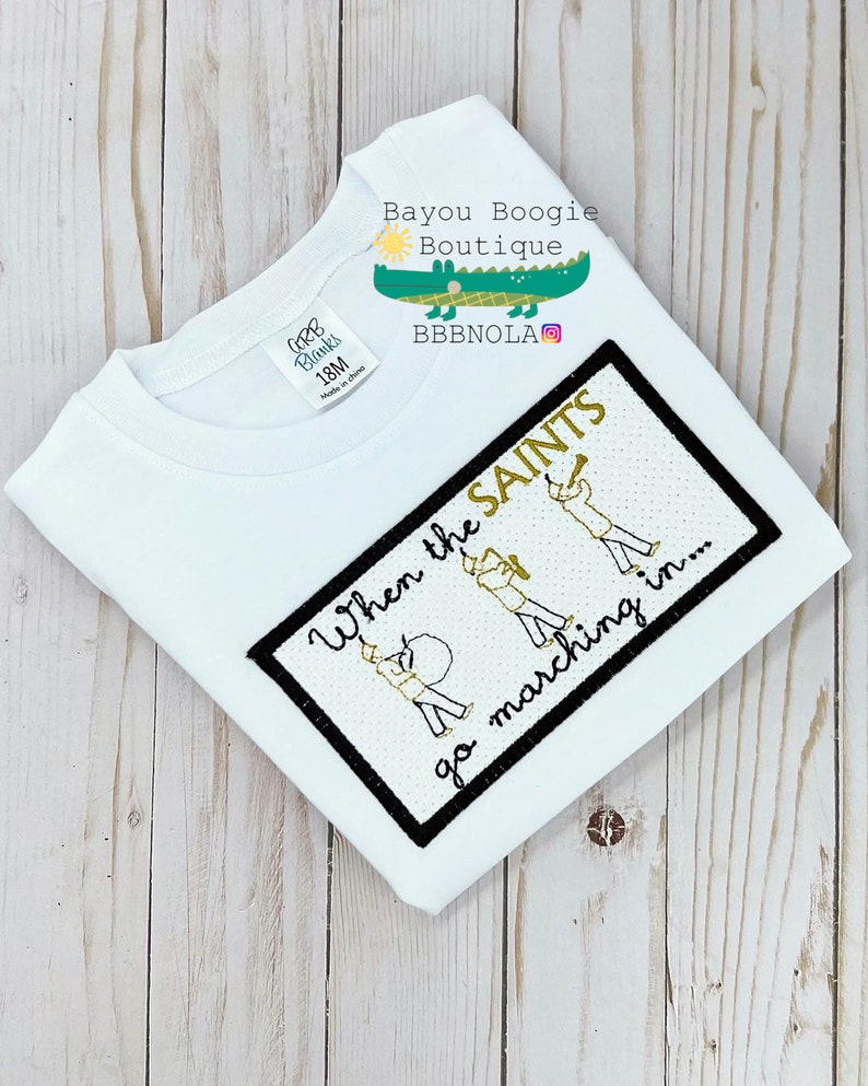 Peut inclure: T-shirt blanc &agrave; manches courtes avec un motif rectangulaire bord&eacute; de noir. Le motif pr&eacute;sente le texte "When the SAINTS go marching in..." et des illustrations de membres d'un groupe de musique. Le t-shirt porte une &eacute;tiquette "CORB Blanks 18M Made in china".