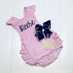 May include: Pink and white striped romper with ruffled trim and the name "Reese" embroidered in navy blue. A large navy blue bow with white trim accents the romper. The romper has snap closures.