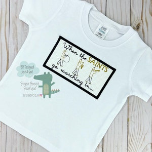 Peut inclure: T-shirt blanc &agrave; manches courtes avec un motif brod&eacute;. Le motif comprend le texte "When the SAINTS go marching in..." et un alligator de dessin anim&eacute;. Le t-shirt porte une &eacute;tiquette indiquant "CARB Blanks 18M".