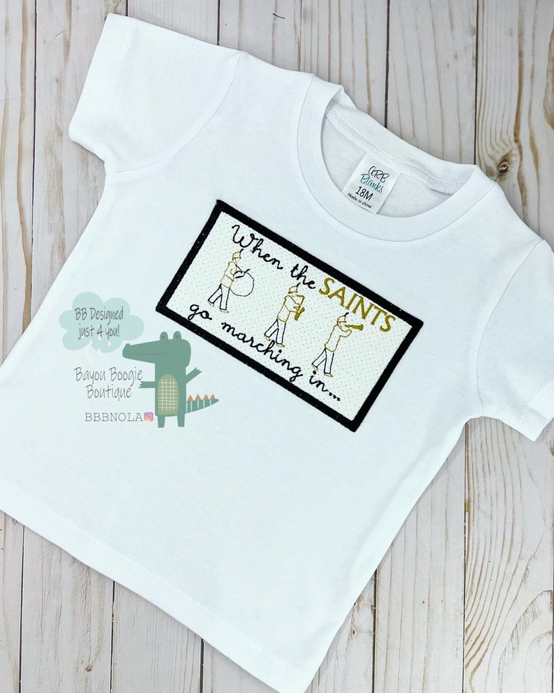Peut inclure: T-shirt blanc &agrave; manches courtes avec un motif brod&eacute;. Le motif comprend le texte "When the SAINTS go marching in..." et un alligator de dessin anim&eacute;. Le t-shirt porte une &eacute;tiquette indiquant "CARB Blanks 18M".