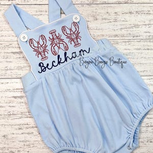 May include: Light blue baby romper with embroidered red lobsters and the name "Beckham" in navy blue script. The romper has white trim, button details, and the text "Bayou Boogie Boutique".
