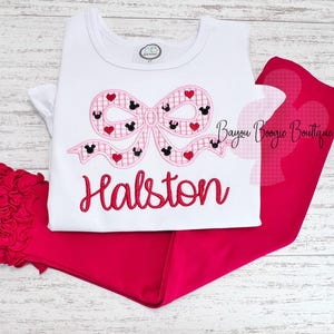 May include: A white t-shirt with a pink bow featuring Mickey Mouse heads and hearts, paired with red pants. The name "Halston" is embroidered in red script. A red ruffled accessory is visible.