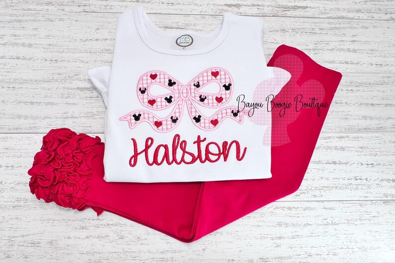 May include: A white t-shirt with a pink bow featuring Mickey Mouse heads and hearts, paired with red pants. The name "Halston" is embroidered in red script. A red ruffled accessory is visible.