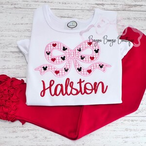 May include: A white t-shirt and red pants set. The shirt features a pink bow design with Mickey Mouse silhouettes and hearts. The name "Halston" is embroidered in red script below the bow. A red hair accessory is also included.