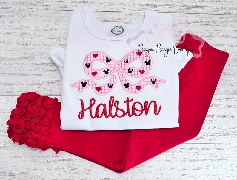 May include: A white t-shirt and red pants set. The shirt features a pink bow design with Mickey Mouse silhouettes and hearts. The name "Halston" is embroidered in red script below the bow. A red hair accessory is also included.