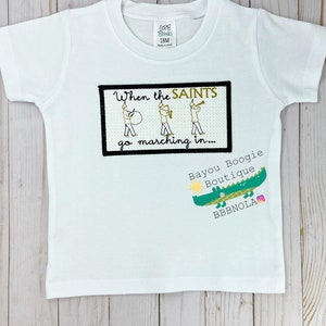 Peut inclure: T-shirt blanc &agrave; manches courtes avec le texte "When the SAINTS go marching in..." et un dessin de trois musiciens. Un logo d'alligator vert avec le texte "Bayou Boogie Boutique" est &eacute;galement visible.