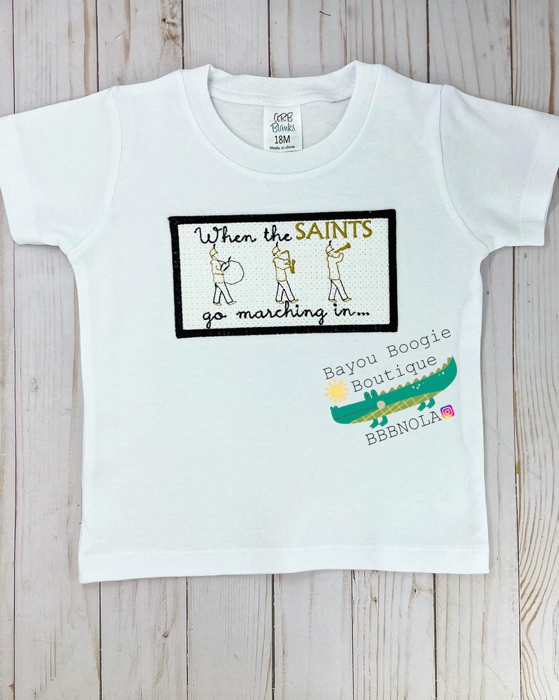Peut inclure: T-shirt blanc &agrave; manches courtes avec le texte "When the SAINTS go marching in..." et un dessin de trois musiciens. Un logo d'alligator vert avec le texte "Bayou Boogie Boutique" est &eacute;galement visible.
