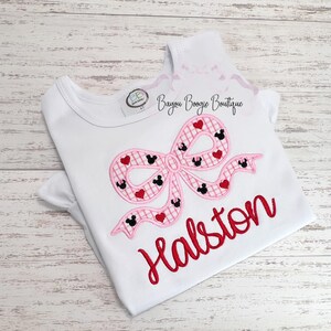 May include: White t-shirt with a pink bow design featuring Mickey Mouse heads and red hearts. The name "Halston" is embroidered in red script below the bow. The shirt has short sleeves and a round neckline. The text "Bayou Boogie Boutique" is printed on the top right.