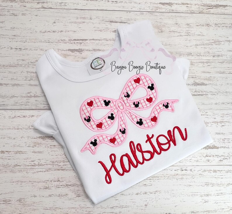 May include: White t-shirt with a pink bow design featuring Mickey Mouse heads and red hearts. The name "Halston" is embroidered in red script below the bow. The shirt has short sleeves and a round neckline. The text "Bayou Boogie Boutique" is printed on the top right.