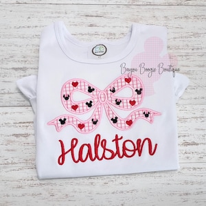 May include: White t-shirt with a pink bow design featuring Mickey Mouse heads and red hearts. The name "Halston" is embroidered in red script below the bow. The shirt has short sleeves and a round neckline.