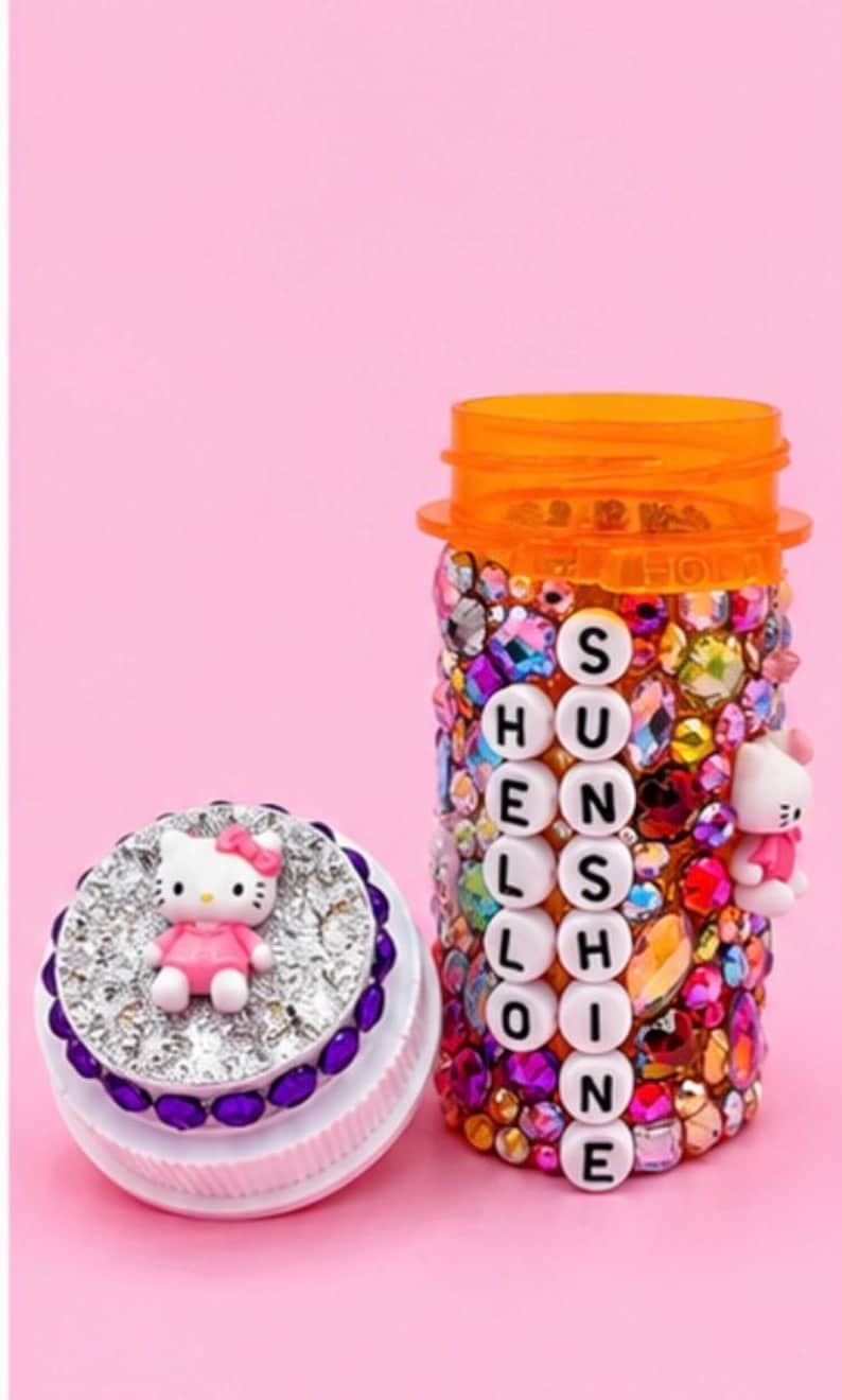 Kawaii-style pill bottle covered in rhinestones with a white cat charm, reading HELLO SUNSHINE