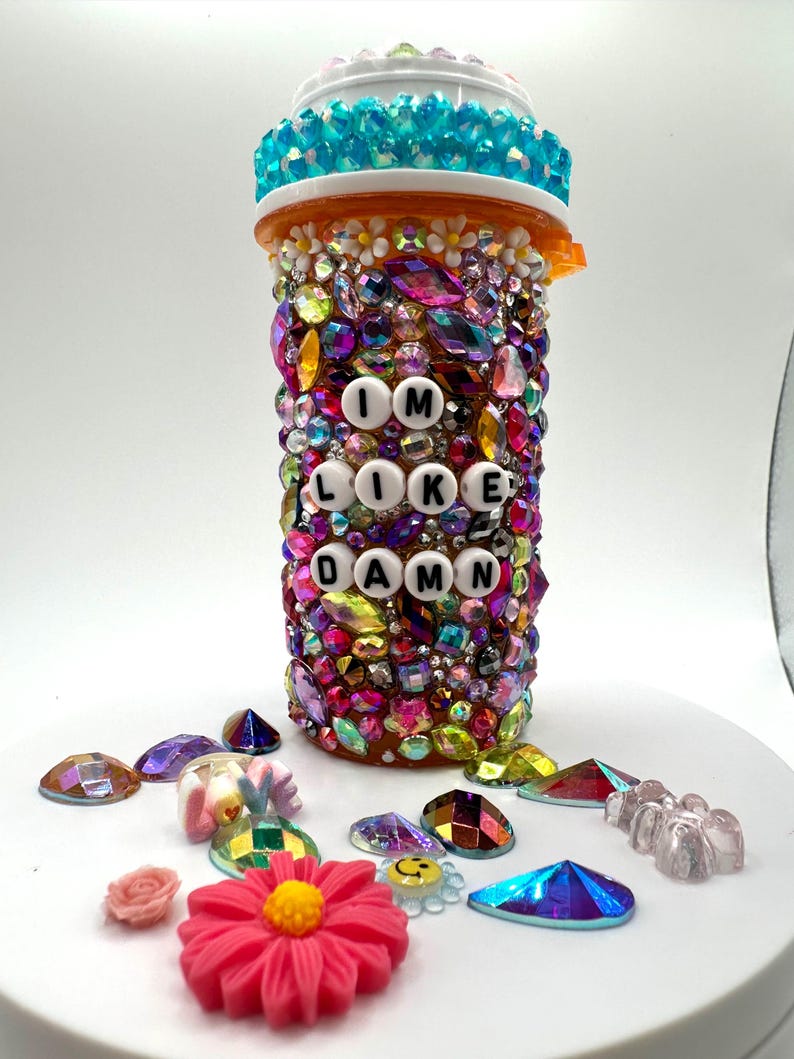 Rhinestone pill bottle covered in multicolor crystals and flower charms, reading I'M LIKE DAMN