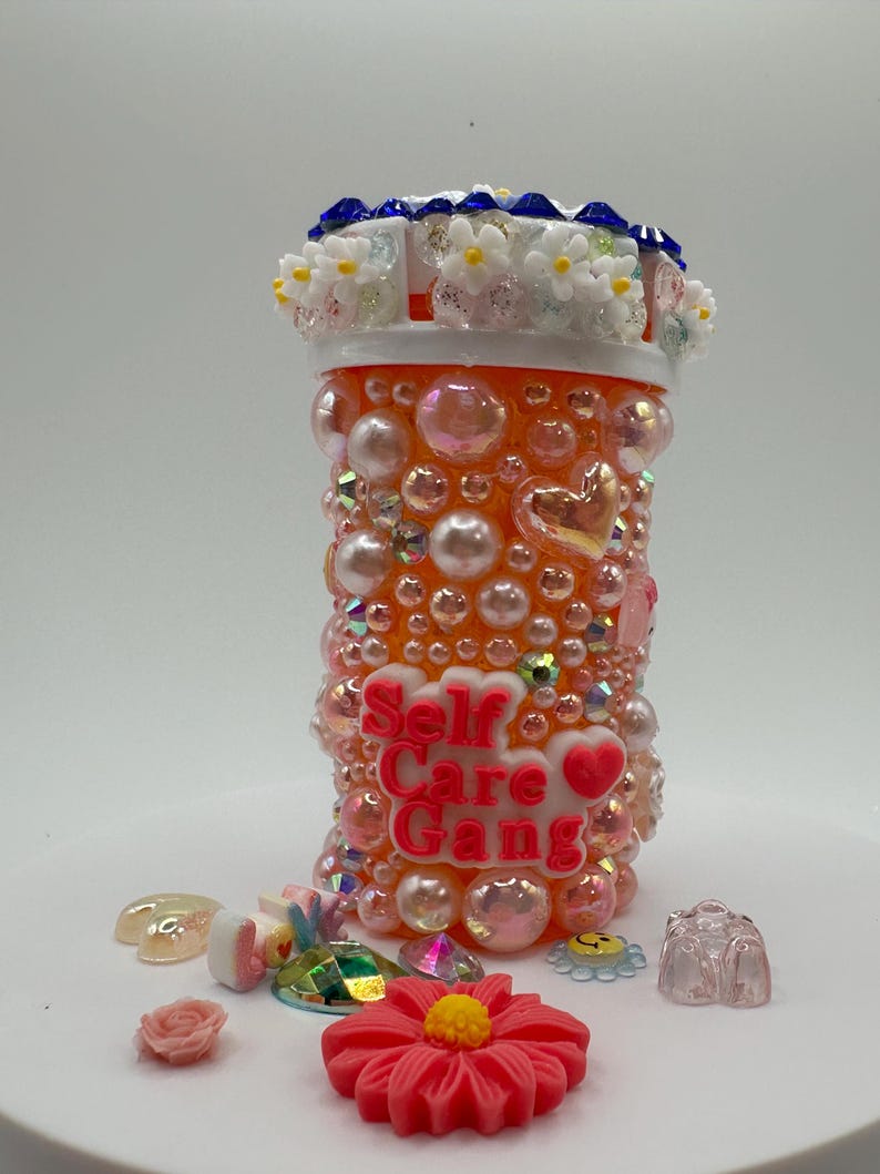 Orange pill bottle decorated with pearls, rhinestones, and flowers, reading Self Care Gang