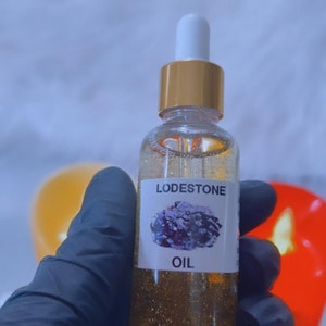 Puede incluir: Una botella de vidrio transparente con tapa dorada y gotero. La botella está llena de un líquido dorado y tiene una etiqueta que dice "Lodestone Oil" con una imagen de una roca gris oscura.