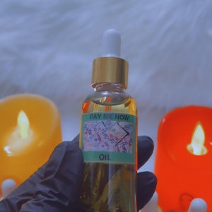 以下が含まれることがあります： 緑色のラベルに「Pay Me Now Oil」と書かれたガラス瓶に入ったオイル。瓶は黒い手袋をした手で持たれている。背景にはオレンジと黄色の2本のろうそくがある。