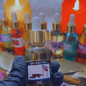 Puede incluir: Una botella de vidrio de aceite con una tapa dorada y una etiqueta que dice "DO AS I SAY" con una ilustración de una mujer de pie sobre un hombre. La botella está llena de un aceite transparente y contiene pequeños objetos redondos blancos. La etiqueta también dice "OIL".