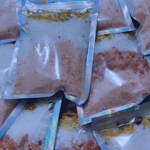 Peut inclure: Gros plan sur plusieurs petits sacs en plastique transparents remplis de sel rose de l'Himalaya et de fleurs séchées. Les sacs sont empilés les uns sur les autres et ont une finition holographique irisée.