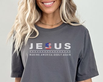 May include: Gray t-shirt with the text "JESUS" in white letters with a small American flag in the "S". Below the text is "MAKING.AMERICA.GODLY.AGAIN" in smaller white letters.
