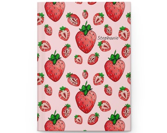May include: A pink notebook with a repeating pattern of red strawberries. The name "Stephanie" is written in black text on the front cover.