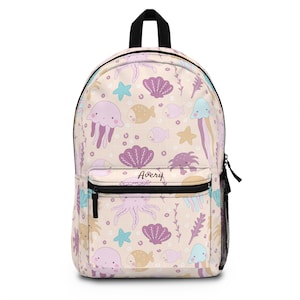 May include: A pink and beige backpack with a sea-themed pattern. The pattern includes jellyfish, starfish, seashells, and fish. The backpack has a black zipper and a black name tag that says "Avery".