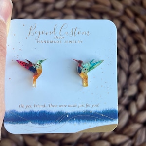 May include: A pair of hummingbird earrings made of resin with a colorful, detailed design. The earrings are on a white card with the text "Beyond Custom Decor HANDMADE JEWELRY" and "Oh yes, Friend... These were made just for you!"