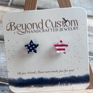 May include: A pair of star-shaped stud earrings on a white card. One earring is navy blue with white stars, and the other is red with white stripes. The card reads "Beyond Custom HANDCRAFTED JEWELRY."