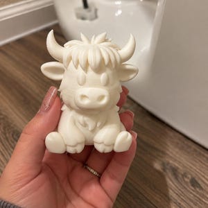 May include: A small, off-white, 3D-printed figurine of a cartoon-style highland cow. The creature features prominent curved horns, a tuft of hair on its head, large oval eyes, and a rounded snout, depicted in a seated position with its front paws forward.