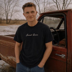 Könnte beinhalten: Schwarzes T-Shirt mit dem Text "Josiah Queen" in Schreibschrift. Die Person trägt das T-Shirt neben einem roten Lastwagen. Der Hintergrund zeigt Bäume und ein Feld.