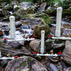 May include: A collection of white PVC pipe water filtration systems with red and brass valves, set among rocks in a stream. Each system has a vertical pipe and horizontal pipes with valves. The background shows a flowing stream.