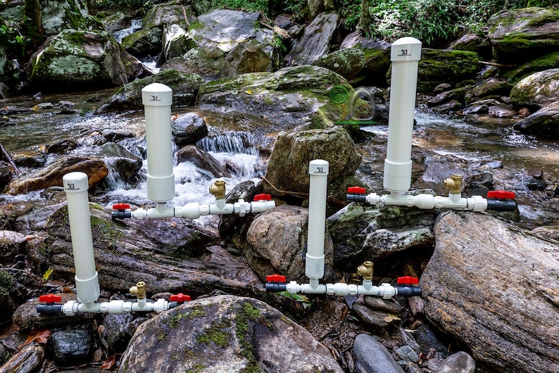 May include: A collection of white PVC pipe water filtration systems with red and brass valves, set among rocks in a stream. Each system has a vertical pipe and horizontal pipes with valves. The background shows a flowing stream.